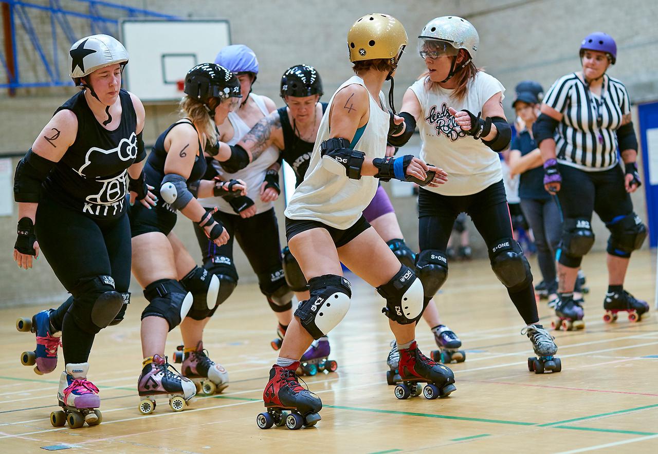 Action from the first All In: Community Roller Derby Short Track scrimmage: from right to left, referree observing (out of focus) from inside of track; paired white team blockers turned slightly to look back at; black team jammer approaching from outside line, pushing gently off from pair of black team blockers, cooperatively stopping white team jammer (obscured by blockers visually).