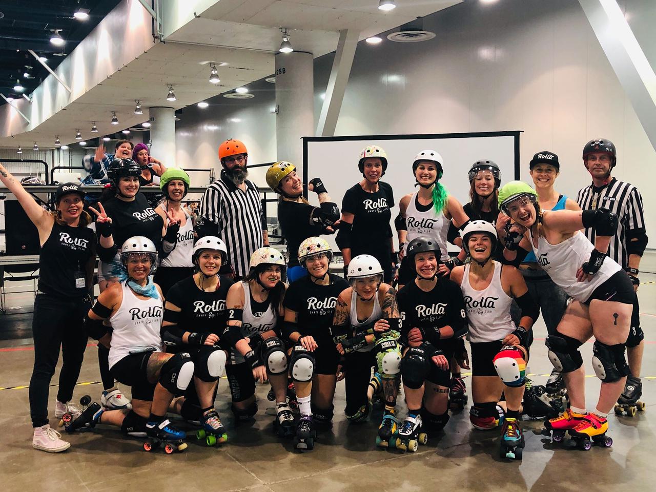 Team photo of the RollaCon 2019 "All Stars" bout roster, and officials. Two rows, front row on one knee. In the background, two people are waving at the camera.