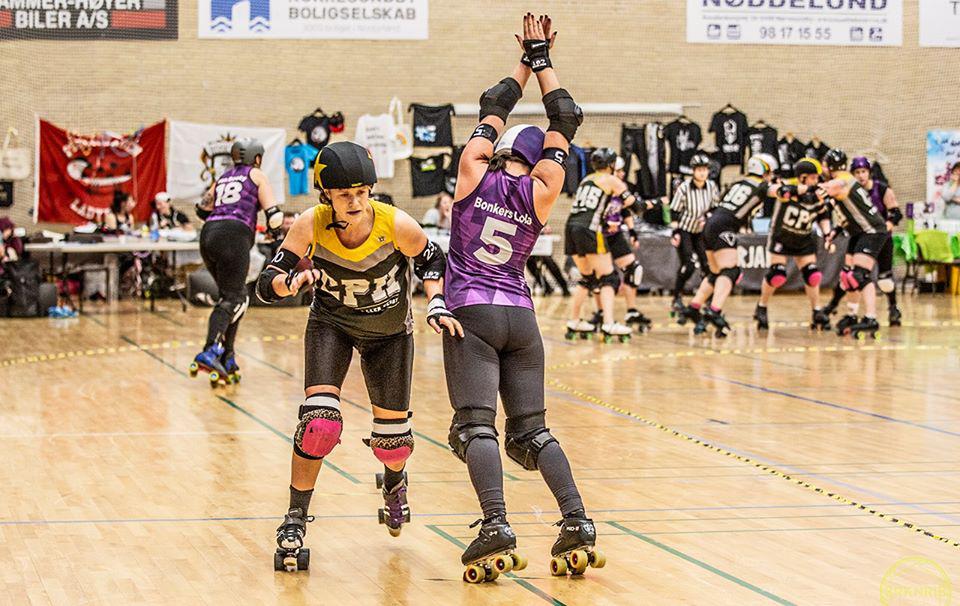 Aalborg (in purple with triangle pattern) versus Copenhagen (in black and yellow), Copenhagen's jammer just getting past lead blocker from Aalborg as she goes out of play.