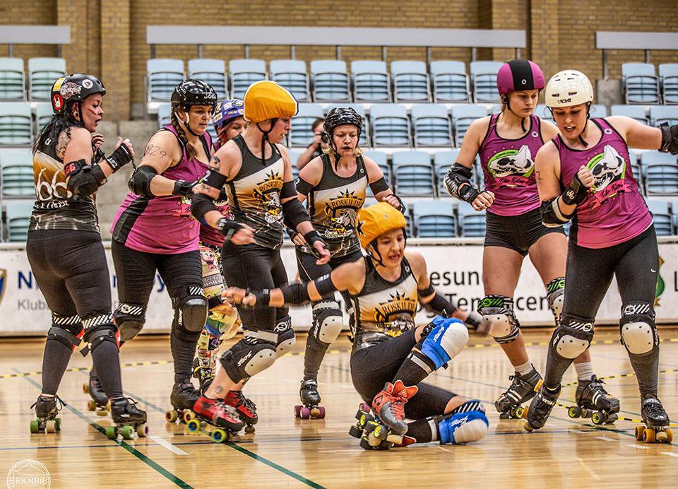 Roskilde (in golden and black) versus O-Town (in purple), dramatic shot with Roskilde's jammer mid-fall after hit from O-Town blocker. Other skaters arrayed around them.